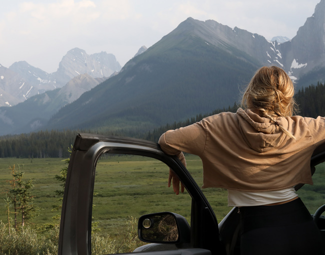 A young woman leaning against her car and looking at the mountains